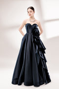 Bathed in soft light, a woman dazzles in a black strapless ball gown. It features a sweetheart neckline, a large ruffled floral detail on the side, and a voluminous skirt. The elegant design creates a sophisticated and glamorous look, perfect for formal events like galas or weddings.1