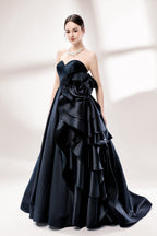 Bathed in soft light, a woman dazzles in a black strapless ball gown. It features a sweetheart neckline, a large ruffled floral detail on the side, and a voluminous skirt. The elegant design creates a sophisticated and glamorous look, perfect for formal events like galas or weddings.2