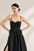 A woman stands against a light background, wearing a black satin gown. The bodice features thin straps, a draped neckline, and beaded embroidery. A high side slit adds allure, combining elegance and sexiness. Perfect for formal events like galas or evening parties, exuding sophistication.6