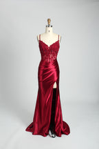 A photograph of a slender, light-skinned woman with long, brown hair in an updo, wearing a red, floor-length evening gown with a deep V-neckline and spaghetti straps. The gown features intricate lace detailing on the bodice and a high slit on the left leg. She accessorizes with a gold ankle bracelet and strappy high heels. The background is a plain, light beige wall with soft shadows cast from a window, creating a minimalist and elegant atmosphere.