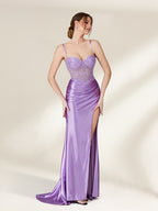 A photograph of a young woman with light skin and brown hair styled in an elegant updo, wearing a shimmering, floor-length, lavender satin gown with a high slit and intricate lace detailing. She stands confidently against a plain, beige background, exuding sophistication and grace.32738-03