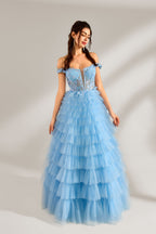 Photograph of a young woman with light skin and dark brown hair styled in loose waves. She wears a voluminous, light blue, multi-layered tulle gown with off-the-shoulder sleeves, intricate lace detailing on the bodice, and a fitted waist. She stands against a plain white background with soft shadows.3