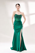 Photograph of a young woman with fair skin and dark hair styled in an elegant updo. She wears a stunning, floor-length, strapless emerald green gown with intricate lace detailing on the bodice and a high slit on the right leg. The dress features a fitted bodice and a flowing, ruched skirt. She accessorizes with large hoop earrings. The background is a soft, white studio setting with diffused lighting, creating a clean, minimalist atmosphere. The woman stands confidently, hands on her hips, exuding grace and