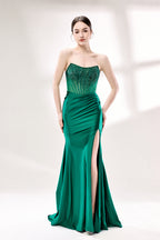 Photograph of a young woman with fair skin and dark hair styled in an elegant updo. She wears a stunning, floor-length, strapless emerald green gown with intricate lace detailing on the bodice and a high slit on the right leg. The dress features a fitted bodice and a flowing, ruched skirt. She accessorizes with large hoop earrings. The background is a soft, white studio setting with diffused lighting, creating a clean, minimalist atmosphere. The woman stands confidently, hands on her hips, exuding grace and