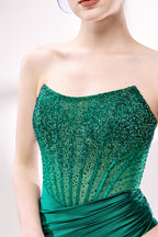 This photograph showcases a close-up of a woman's torso and upper chest. She is wearing a strapless, form-fitting, emerald green dress adorned with intricate beadwork and sequins that shimmer in the light. The dress has a sheer, ruched texture that accentuates her slender figure. Her skin is fair, and her lips are painted a soft pink. The background is a plain, light grey, ensuring that the vibrant green of her dress stands out prominently. The image has a professional, high-fashion feel, with a focus on el