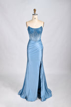 Photograph of a mannequin wearing a strapless, floor-length, royal blue evening gown. The gown features intricate beadwork and embroidery along the bodice, transitioning into a smooth, flowing fabric. The dress has a high slit on the left side, adding a modern touch. The mannequin stands against a plain, light gray background, emphasizing the elegance and sophistication of the gown. The mannequin's head is not visible.