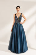 A photograph of a woman in an elegant, floor-length, sleeveless gown. The dress features a deep V-neck adorned with intricate beadwork and sequins in shades of blue, green, and gold. The bodice is fitted, transitioning into a voluminous, blue tulle skirt. Her dark hair is styled in a sleek bun, and she wears gold earrings. The background is a plain, light beige, allowing the dress's details to stand out.