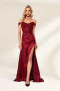 Photograph of a slender, attractive blonde woman with wavy hair. She wears a floor-length, off-shoulder, burgundy satin gown with a high slit and intricate beaded bodice. Her makeup is glamorous, with bold red lipstick. She stands against a plain white background, exuding elegance and sophistication.01