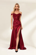 Photograph of a slender, attractive blonde woman with wavy hair. She wears a floor-length, off-shoulder, burgundy satin gown with a high slit and intricate beaded bodice. Her makeup is glamorous, with bold red lipstick. She stands against a plain white background, exuding elegance and sophistication.01