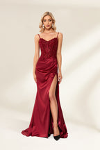 Photograph of a slender, attractive blonde woman with wavy hair. She wears a floor-length, off-shoulder, burgundy satin gown with a high slit and intricate beaded bodice. Her makeup is glamorous, with bold red lipstick. She stands against a plain white background, exuding elegance and sophistication.03