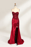 This photograph features a mannequin dressed in a strapless, floor-length, deep red satin evening gown. The gown is adorned with intricate sequin embroidery on the bodice and a high slit on the left side, revealing black stockings. The fabric has a smooth, luxurious texture. The mannequin stands on a black stool against a plain white background, creating a stark contrast that highlights the gown's vibrant color and detailed design. The overall style is elegant and glamorous, suitable for formal occasions.1
