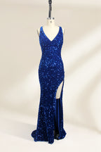 Photograph of a mannequin wearing a floor-length, deep V-neck, navy blue sequin dress with a high slit on the right side. The dress has a fitted bodice and a flared skirt. The background is a plain, light beige. The mannequin stands on a white pedestal, and the dress sparkles under soft, diffused lighting.01