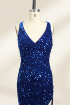 Photograph of a mannequin wearing a floor-length, deep V-neck, navy blue sequin dress with a high slit on the right side. The dress has a fitted bodice and a flared skirt. The background is a plain, light beige. The mannequin stands on a white pedestal, and the dress sparkles under soft, diffused lighting.03
