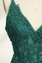 Photograph of a dark green, floor-length evening gown displayed on a white dress form. The gown features a deep V-neckline with thin spaghetti straps, a fitted bodice adorned with intricate lace detailing, and a flowing, slightly flared skirt. The dress showcases a rich, textured fabric with a shimmering effect, accentuating the luxurious and elegant style. The background is a plain, off-white color, ensuring the dress remains the focal point.06
